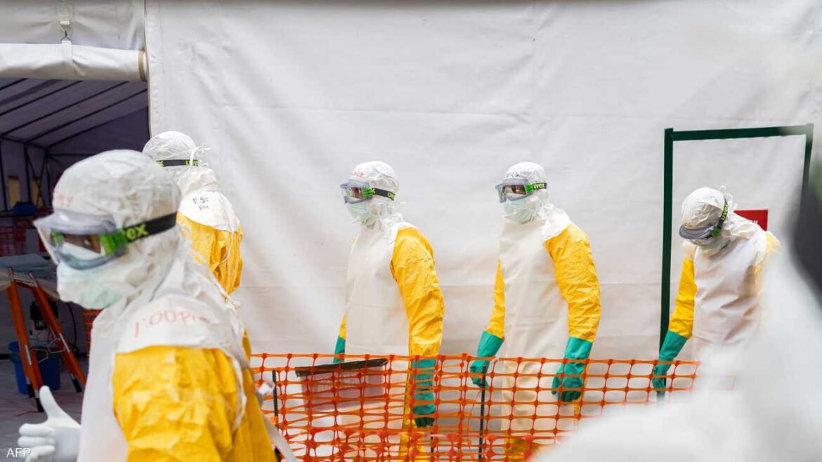 Medical workers in full hazmat suits and goggles stand behind orange safety barrier outside a white tent.