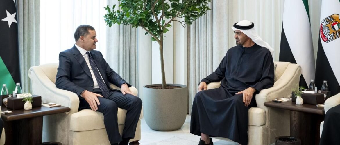 Two men in formal attire sit in beige armchairs, talking in a meeting room with flags in the background.