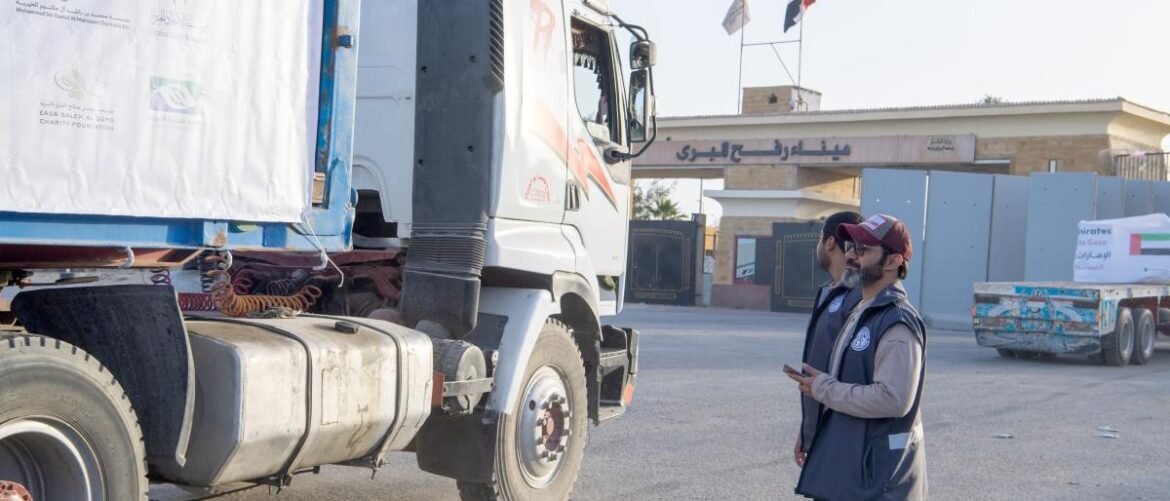 Two aid workers in vests stand beside a large white cargo truck in a depot, with a building and flags in the background.