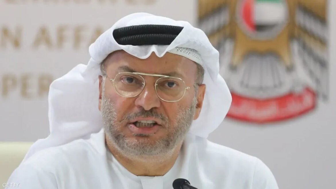 Man in a white kandura and ghutra speaking at a press conference with a blurred emblem in the background; wearing glasses and a short beard.