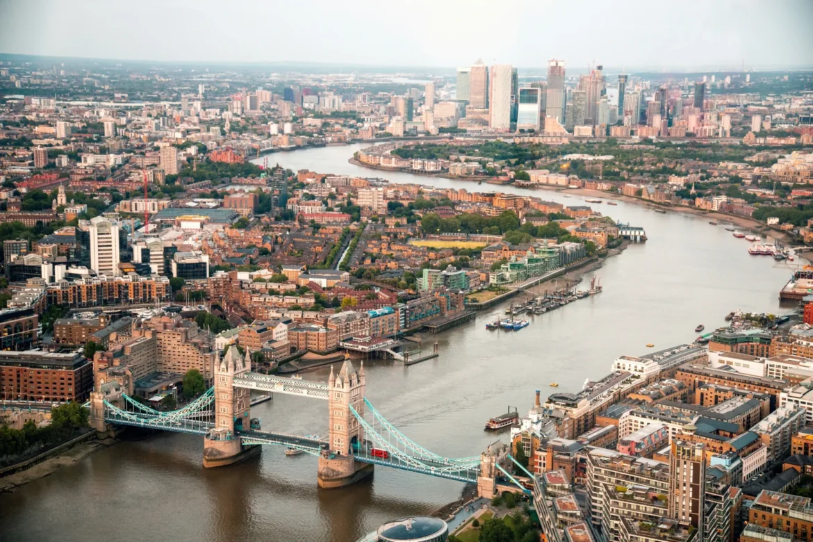 Aerial view of central London with the Thames River and Tower Bridge in the foreground, and a dense city skyline in the distance.