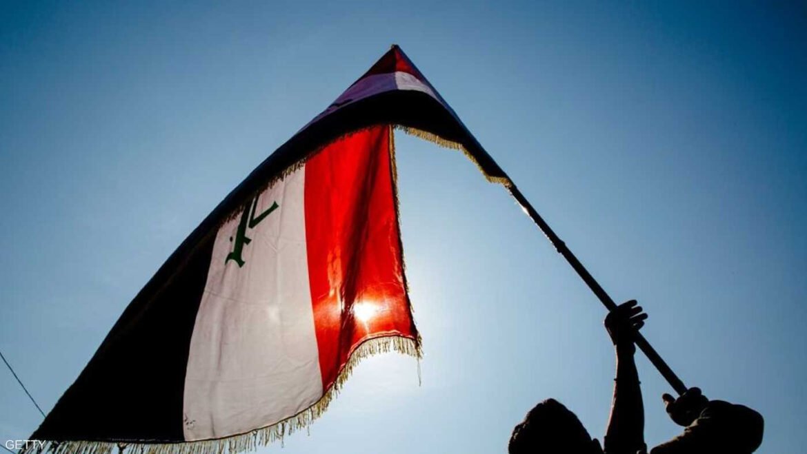Silhouette of a person raising an Iraqi flag against a bright blue sky behind the flagpole.