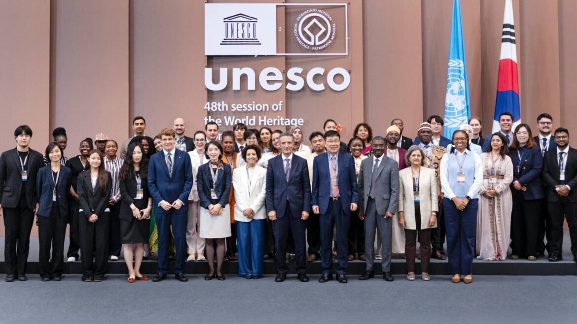 Group of diverse delegates posing for a group photo in front of UNESCO signage at the 48th World Heritage session. Flags appear in the background on the right.