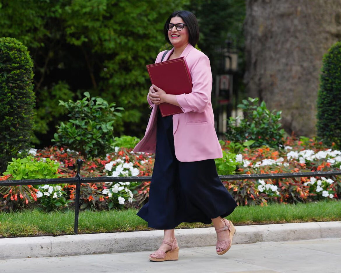 Woman in a pink blazer walks along a sidewalk, holding a red folder.