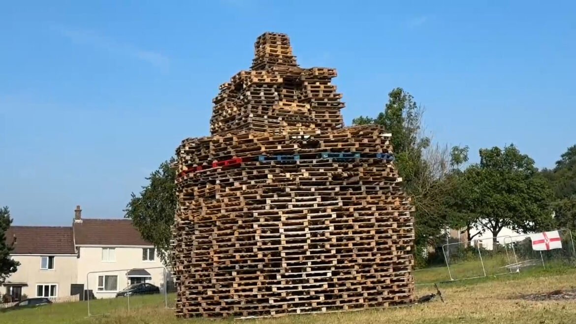 Tall pyramid of stacked wooden pallets in a grassy field with suburban houses nearby and a blue sky behind it.