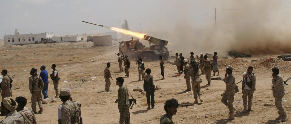 Group of soldiers in desert combat gear watching a rocket launcher firing during a battle, with smoke and vehicles in the background, conveying a war scene.