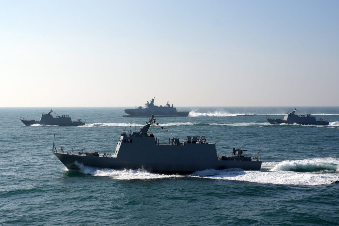 Several gray warships sailing in formation across an open blue sea under a clear sky, with waves at the bow of the foreground ship.”