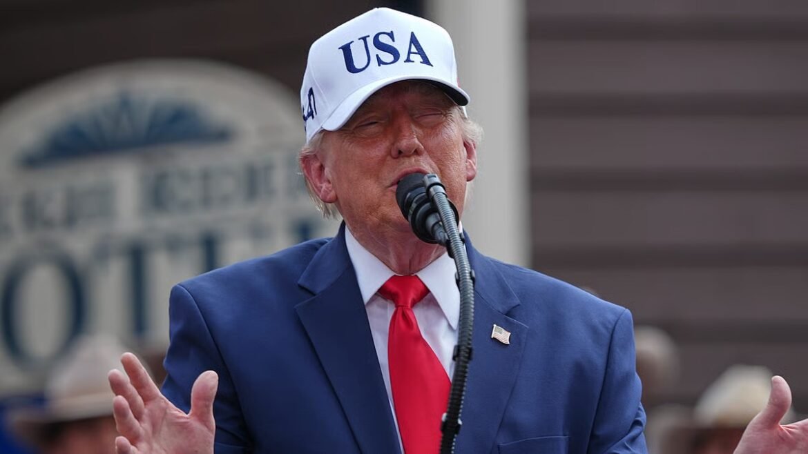 Man in a blue suit and red tie wearing a white USA cap, speaking into a microphone.