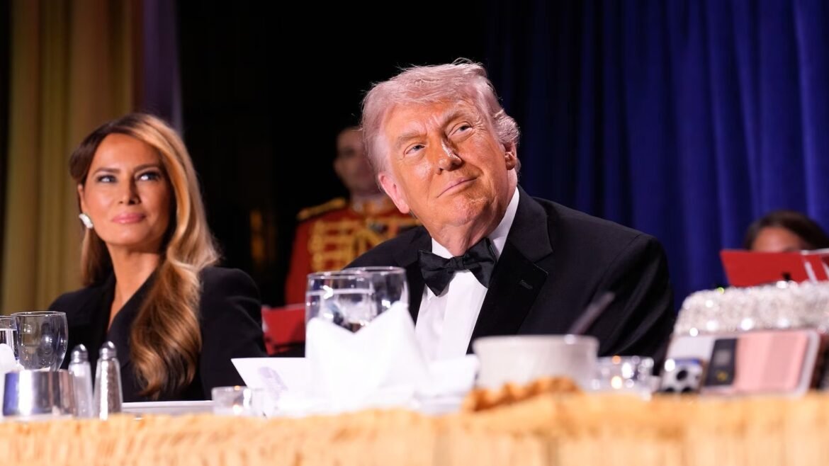 Donald Trump in a black tuxedo sits at a banquet table beside Melania Trump during a formal event, both looking to the side as a stage backdrop looms behind them.