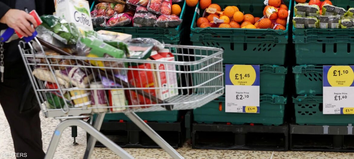 Shopping cart overflowing with groceries in a supermarket, with fruit crates and price signs in the background.