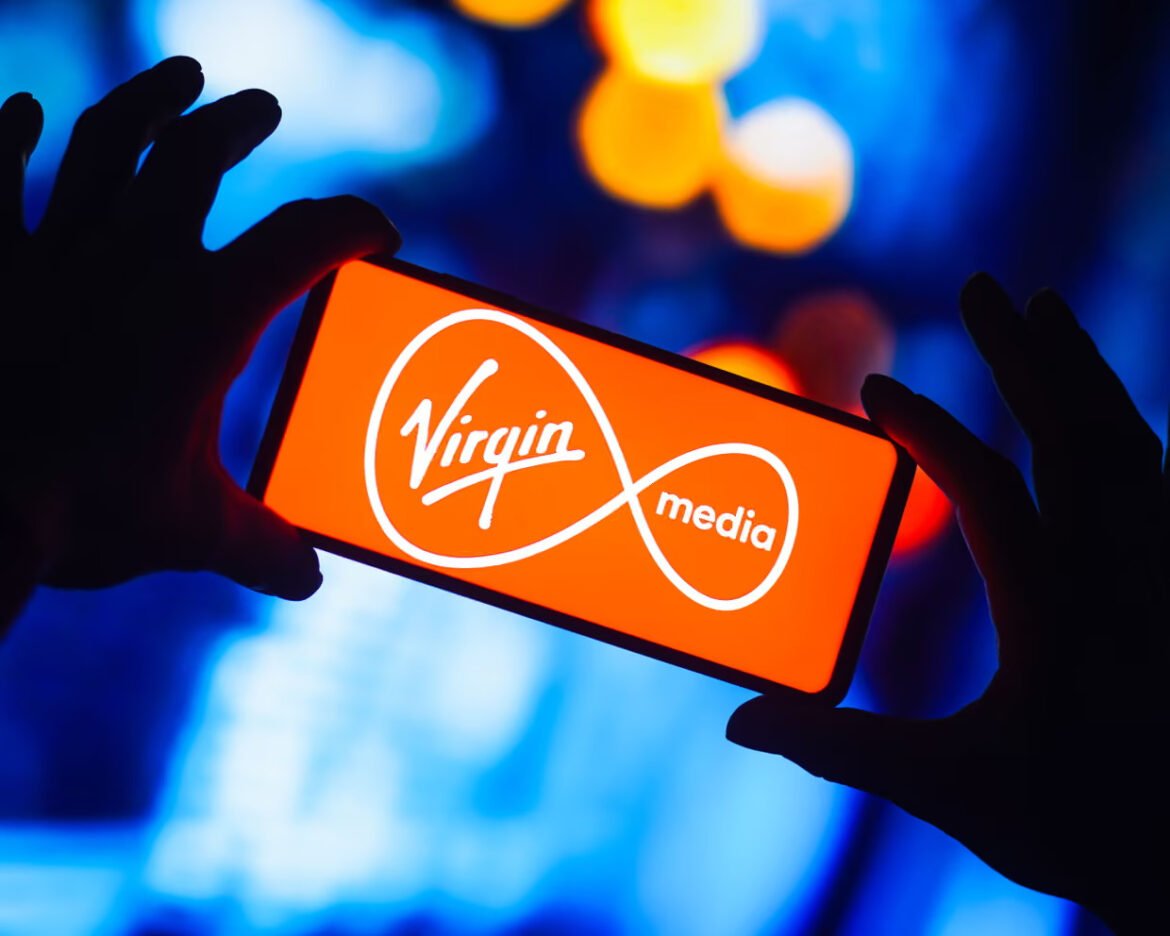 Two hands hold a smartphone displaying the Virgin Media logo on an orange screen.