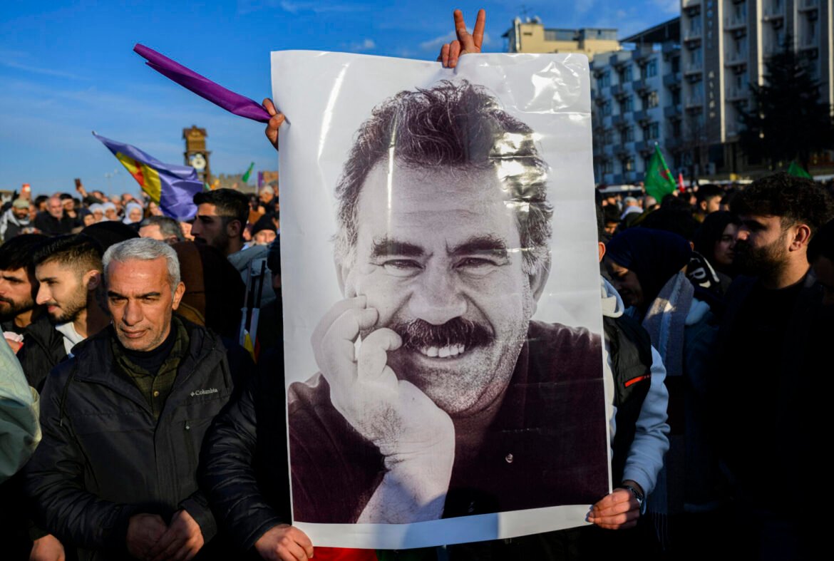 Crowd at a protest holding a large poster of a smiling man against a city street backdrop.