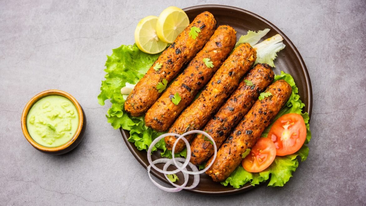 Five grilled kebabs arranged on lettuce with lemon wedges, tomato slices and onion rings, plus a green dipping sauce on the side.