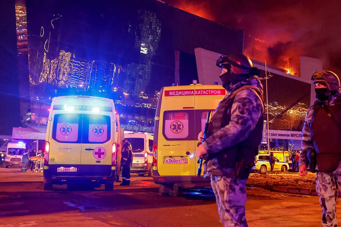 Night scene with two ambulances and armed responders outside a burning building with blue and orange lights flashing at night.