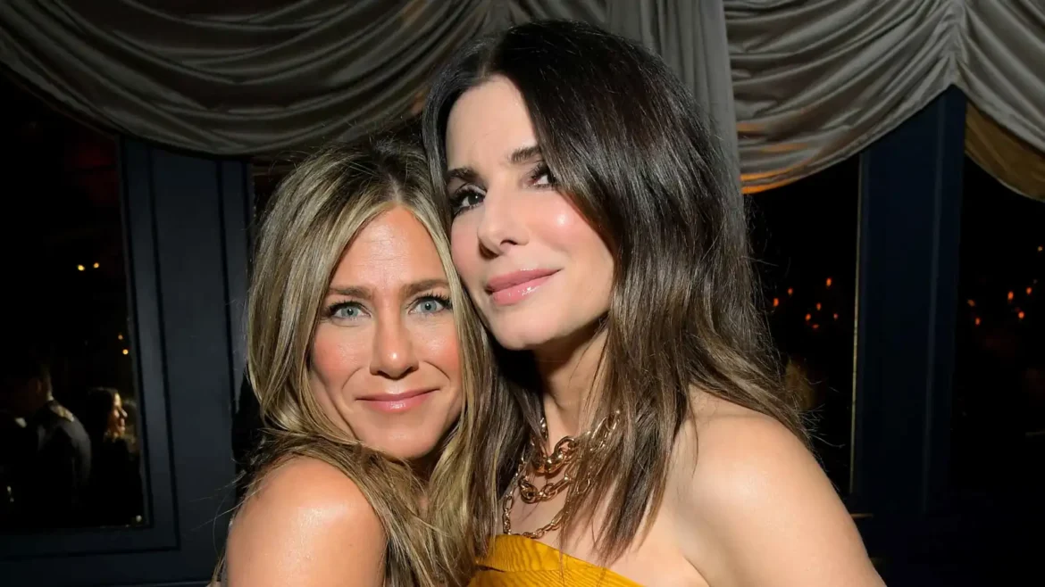 Two women posing closely together and smiling at the camera at a social event, one wearing a yellow outfit and a gold necklace.
