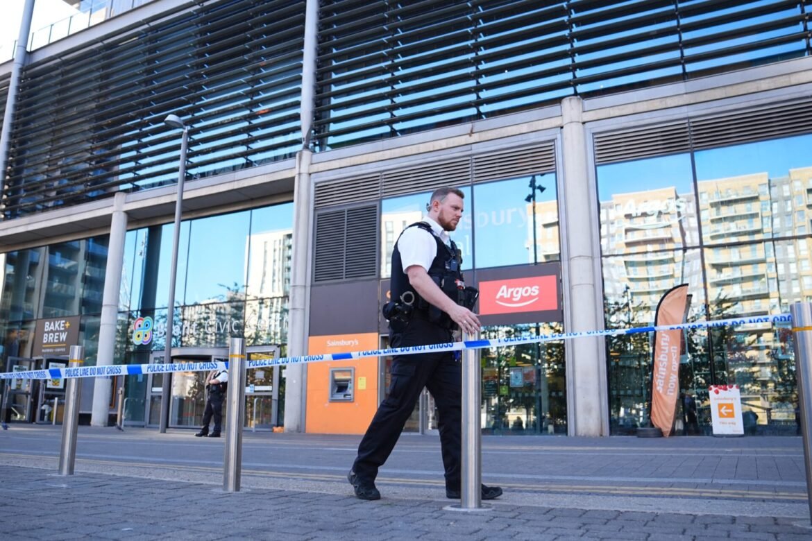 Police officer in tactical vest walking past blue-and-white 'Police line do not cross' tape outside a glass-fronted building district.