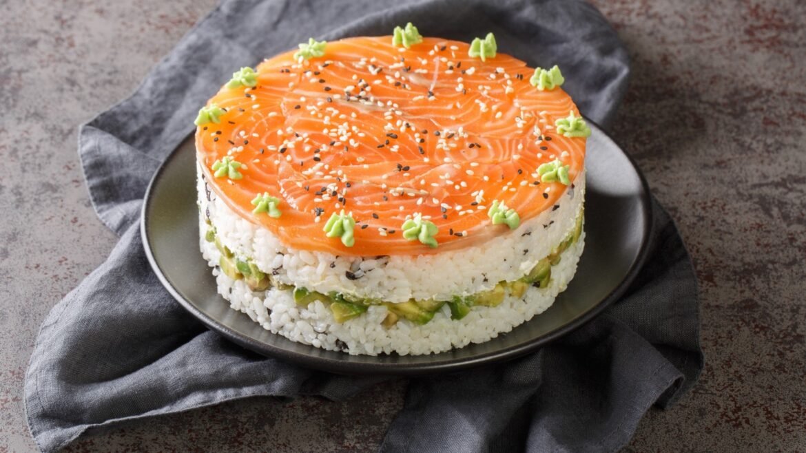 Round sushi-inspired cake: salmon-topped rice layers with avocado bits, sesame seeds, and green wasabi decorations on a dark plate.