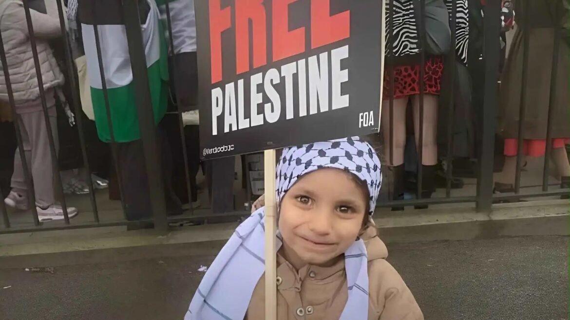 Young child smiles while holding a protest sign that reads 'Free Palestine' in front of a crowd behind a railing.