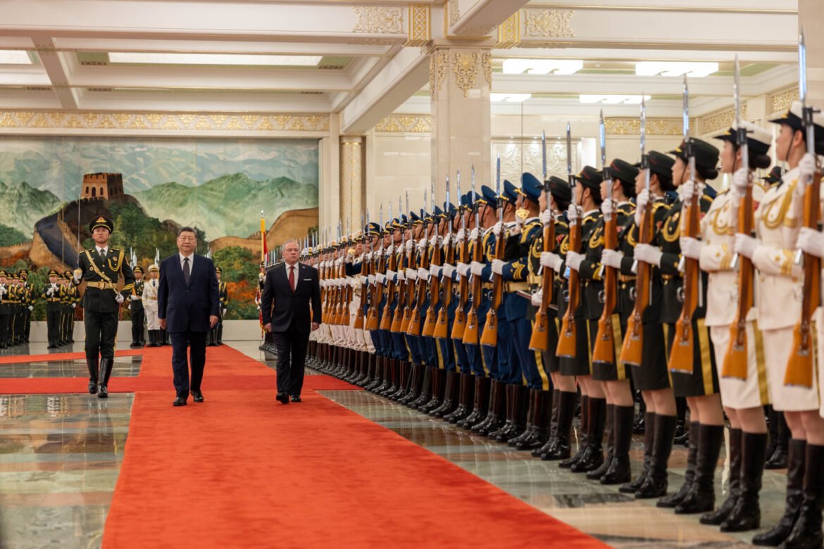 Two men in formal suits walk along a red carpet flanked by rows of uniformed guards with rifles in a grand hall.