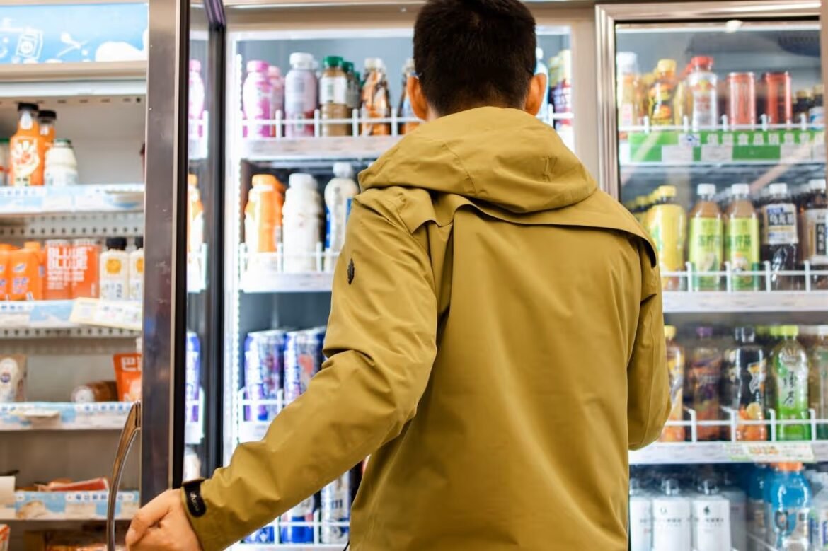 Man in a tan jacket seen from behind selecting drinks from a backlit refrigerated display.