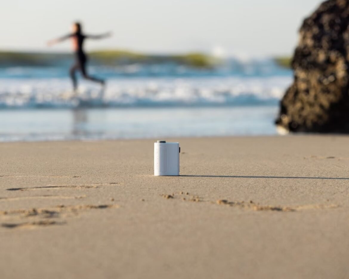 Small white cylindrical container standing upright on sandy beach with a blurred runner and ocean in the background.