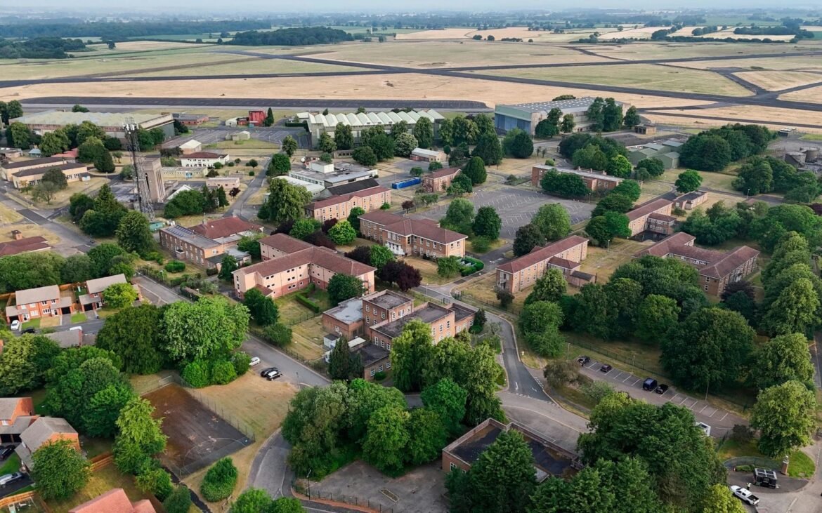 Aerial view of a small town with brick apartment blocks, streets, and many green trees, set against open fields in the distance