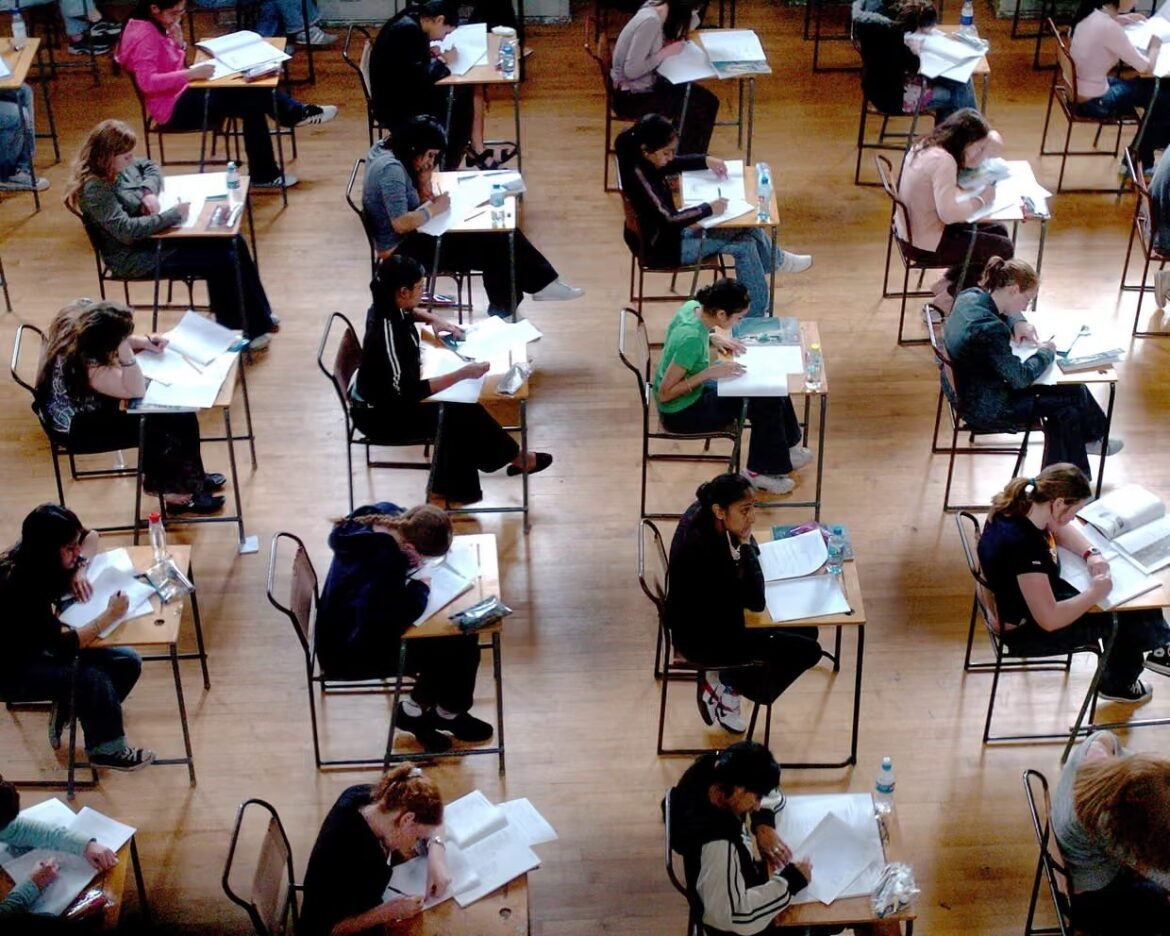 Top-down view of students sitting at individual desks spaced apart in a large room, studying and writing on papers.