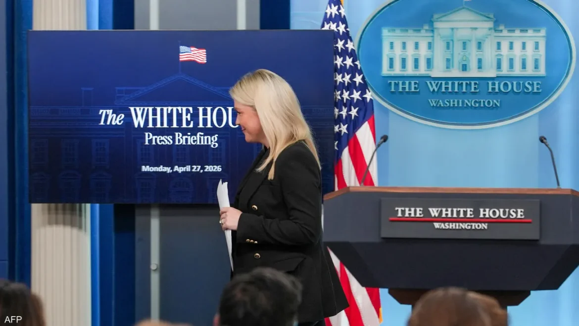 Blonde woman in a black blazer walks with papers past a screen that says The White House Press Briefing, with the podium and American flag nearby.