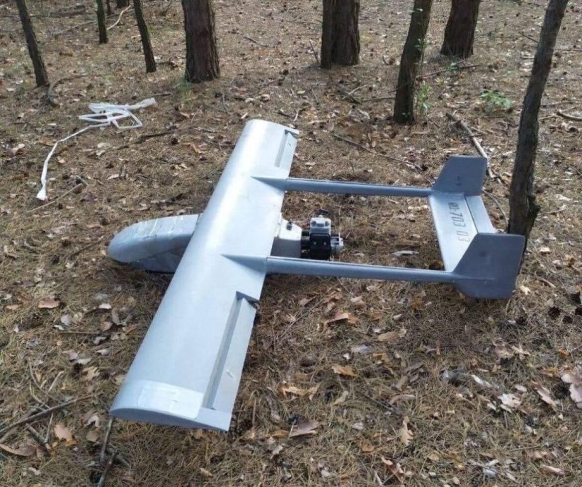 Metal drone/RC aircraft wing lying on pine-needle ground in a forest, with a small engine mount in the center and twin tail sections at the rear.