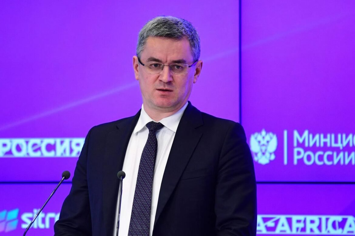 Man in a dark suit and tie speaking at a press conference, standing behind a podium with microphones, purple backdrop with Russian text behind him.