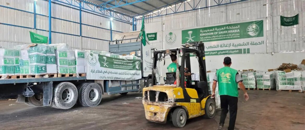 Green bannered warehouse scene with a yellow forklift loading aid pallets onto a truck; banners read Kingdom of Saudi Arabia and King Salman Humanitarian Aid & Relief Centre in Arabic/English (humanitarian operation).