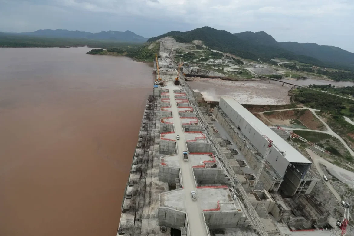 Aerial view of a dam under construction along a muddy brown river, with cranes, concrete spillways, and a long road of activity nearby.