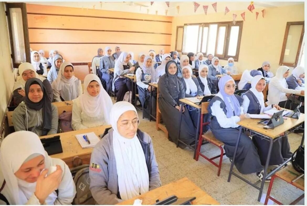 A bright classroom filled with female students wearing hijabs, seated at desks with notebooks and tablets, looking toward the front of the room.