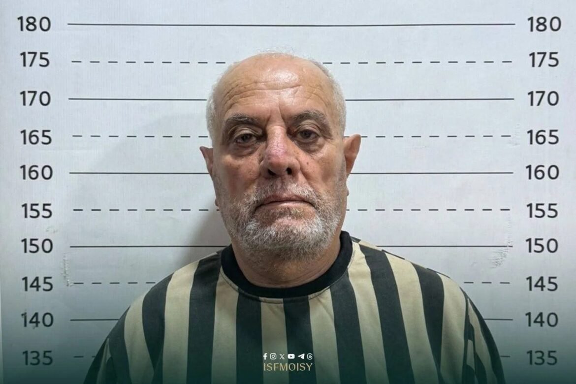 Mugshot photo of an older man wearing a striped prison uniform, standing in front of a height‑measurement wall.