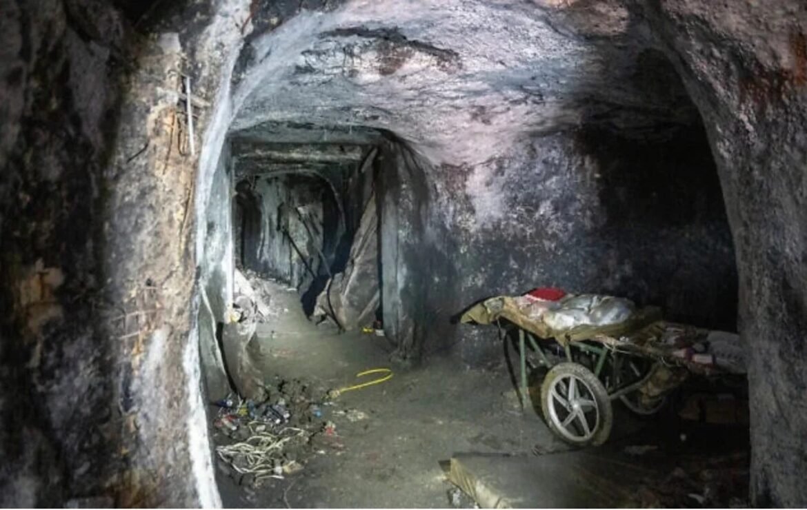Dark underground mining tunnel with a small wagon loaded with supplies along the right wall and rough rock walls surrounding it.