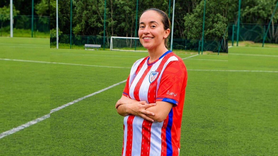 Female soccer player stands with arms crossed on a green training field, wearing a red-and-white striped jersey with a club crest on the chest.