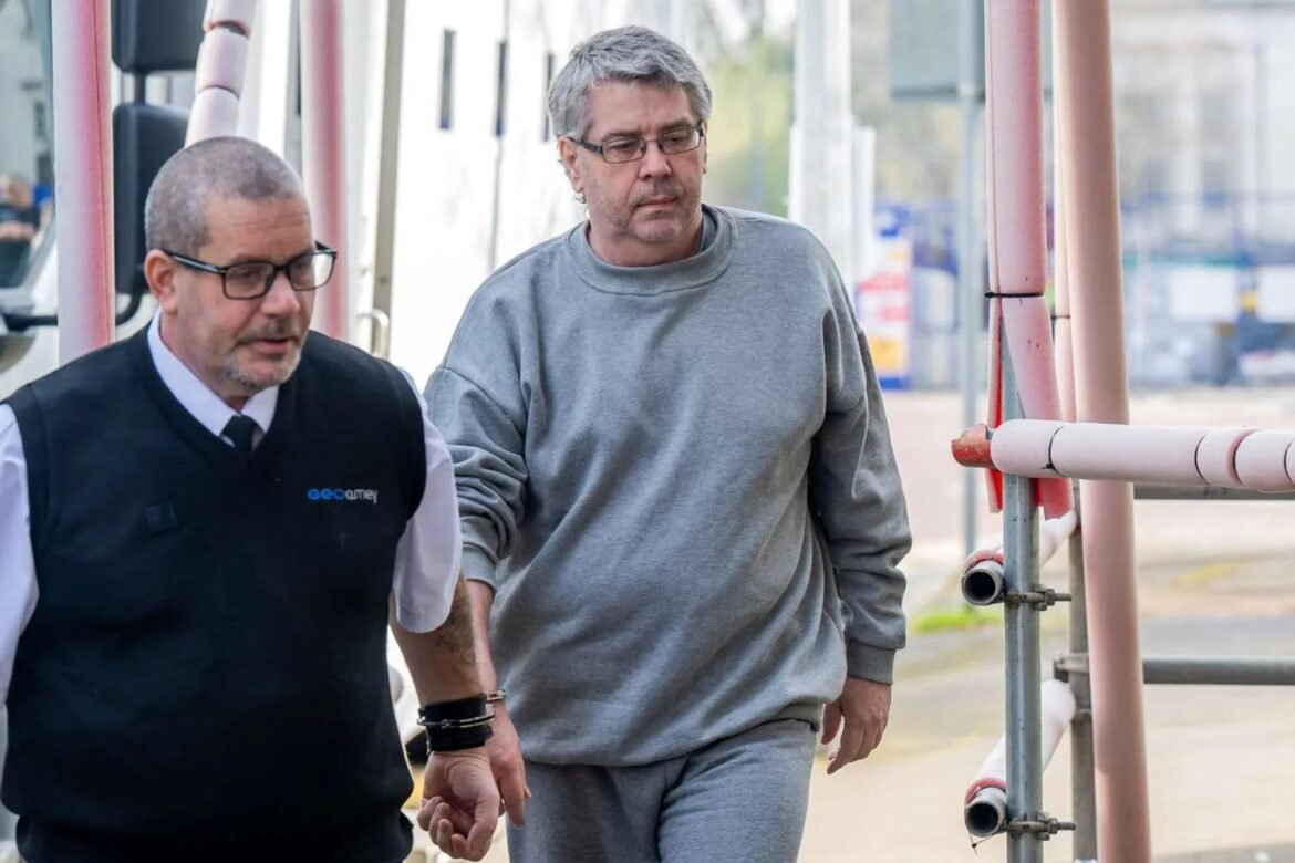 Two men walk on a construction site beside pink scaffolding, one in a gray sweatshirt and the other in a black vest and glasses.