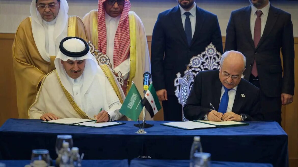 Two men sign official documents at a formal bilateral agreement ceremony: one in a white thobe and head coverings, the other in a dark suit and blue tie, with small Saudi and Syrian flags on the table and officials standing behind.