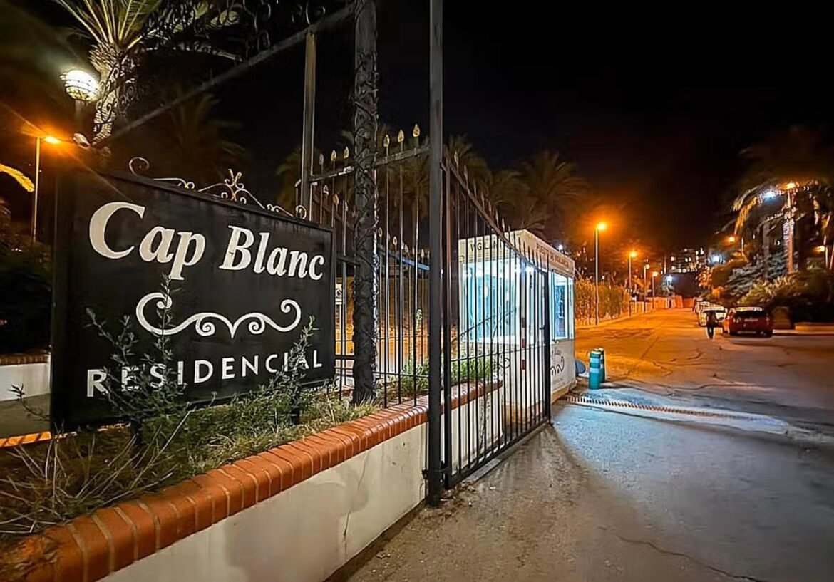 Nighttime view of the Cap Blanc Residencial entrance with a wrought-iron gate and large sign, palm trees in the background, streetlights glowing.