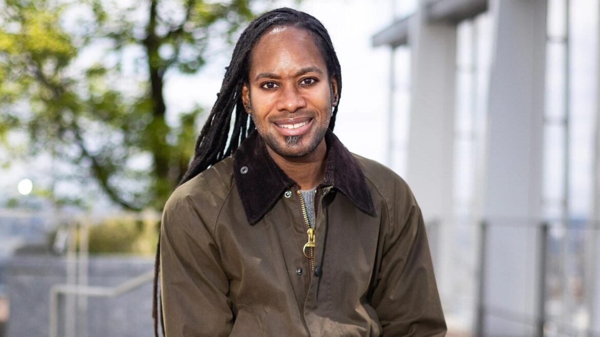 Smiling man with long dreadlocks wearing a brown jacket outdoors, with a modern building in the background.