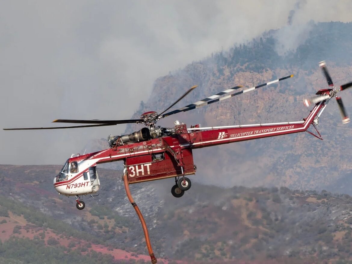 Red firefighting helicopter in flight, with a water bucket suspended from a long hose, against a smoky hillside fire.