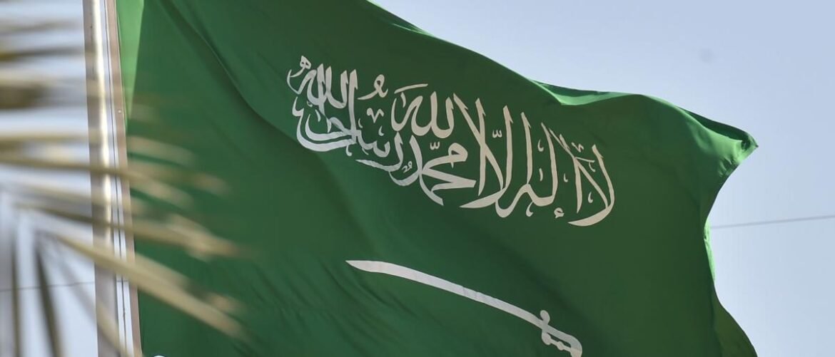 Green Saudi Arabian flag waving in the wind, with white Arabic inscription and a sword symbol beneath it.