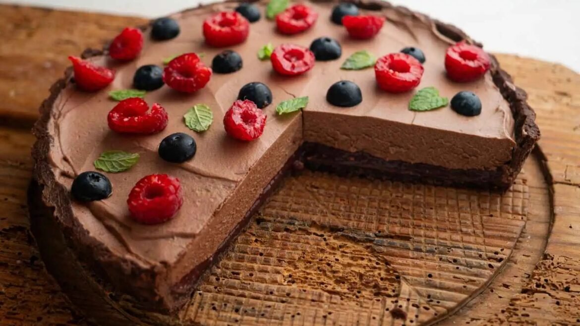 Chocolate cheesecake topped with raspberries, blueberries, and mint on a rustic wooden board, with a slice removed to reveal the creamy filling.