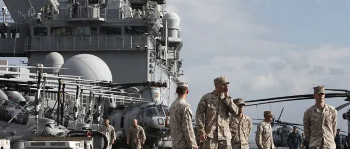 Military personnel in camouflage uniforms walk on the deck of a large aircraft carrier, with helicopters and ship structures in the background.