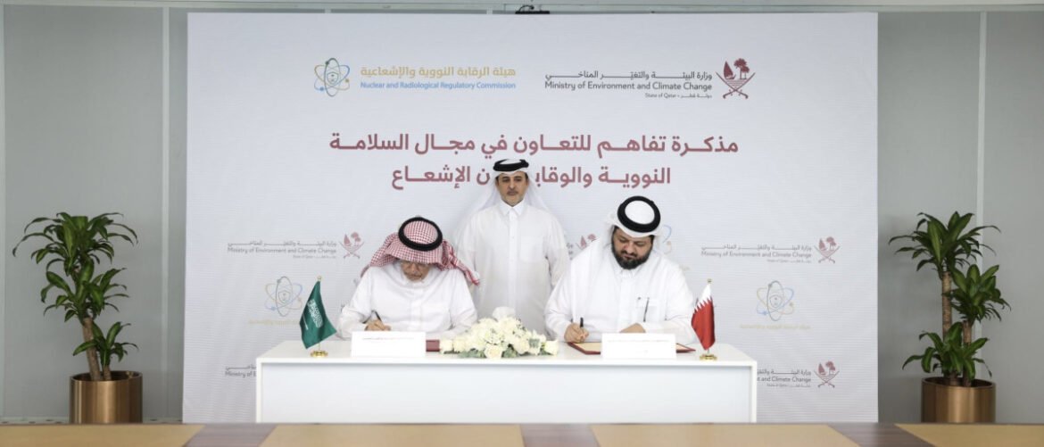 Two men in white thobes sign documents at a formal ceremony, with Saudi and Qatari flags on the table and a standing official behind them.