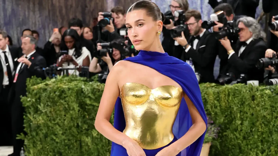Woman on a red carpet wearing a metallic gold strapless corset and a royal blue cape amidst photographers.