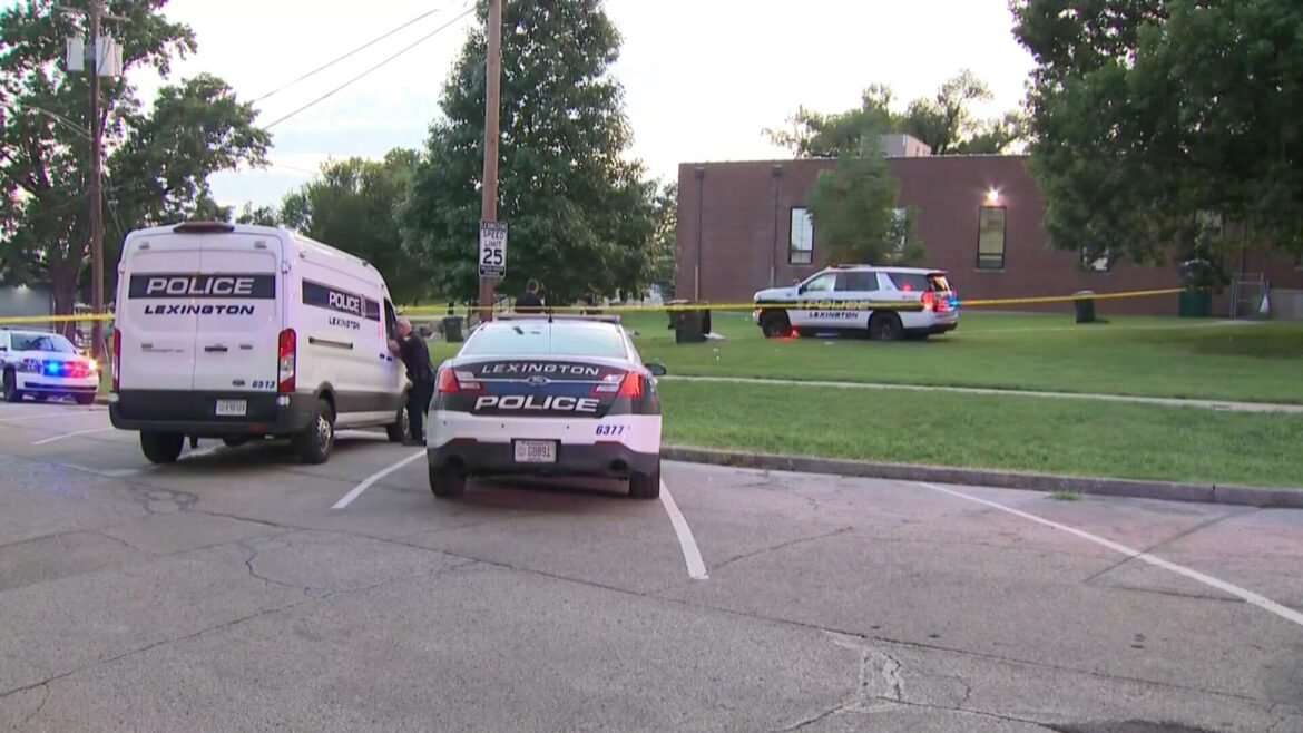 Police vehicles line a street with yellow crime tape around a grassy area near a brick building, scene of an active incident