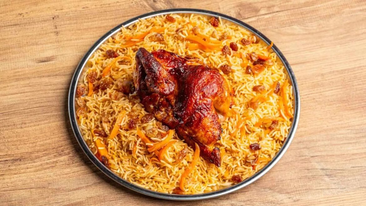 Plate of biryani with a glazed roasted chicken leg on top, scattered carrots and raisins on a wooden table surface