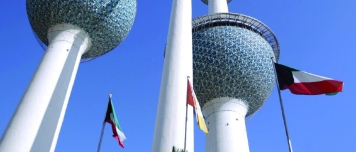 Kuwait Towers with white columns and mosaic spheres, flags waving in front against a clear blue sky.
