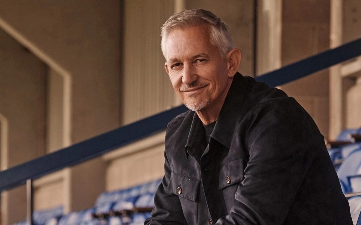 Smiling older man with gray hair in a black jacket leaning on a railing in an indoor stadium stands.